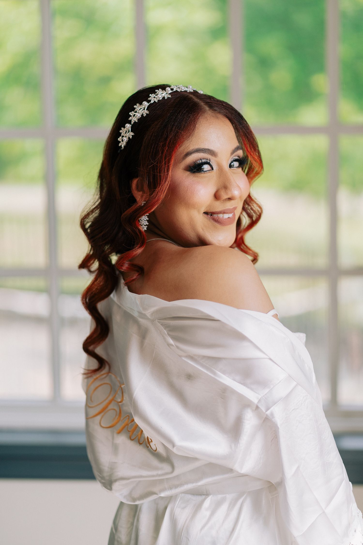 Smiling bride in white satin robe with crystal headband and red curly hair poses near bright window.