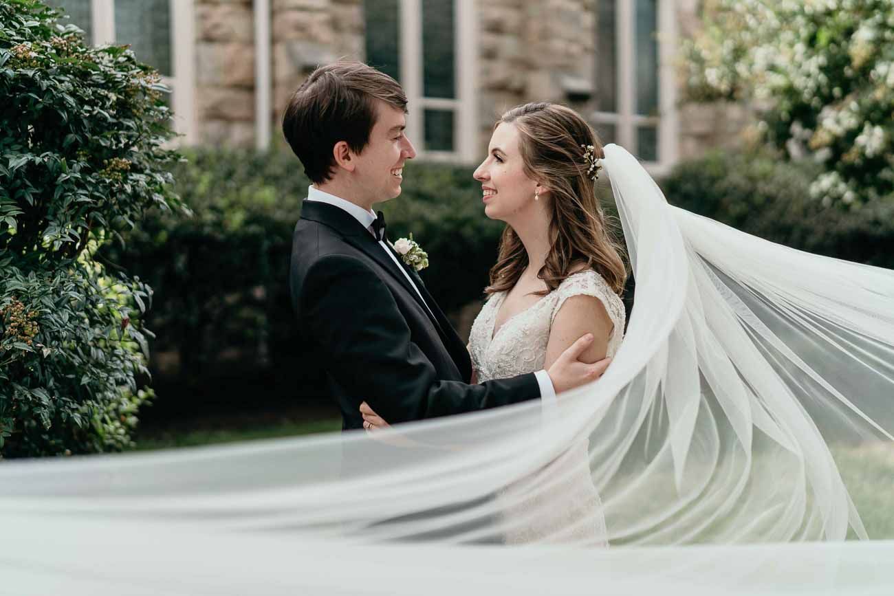 Bride and groom gazing at each other as her veil flows in the wind outside a stone church.