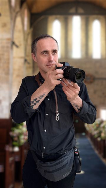 Tattooed male photographer holding a DSLR camera inside a church with pew flowers visible.