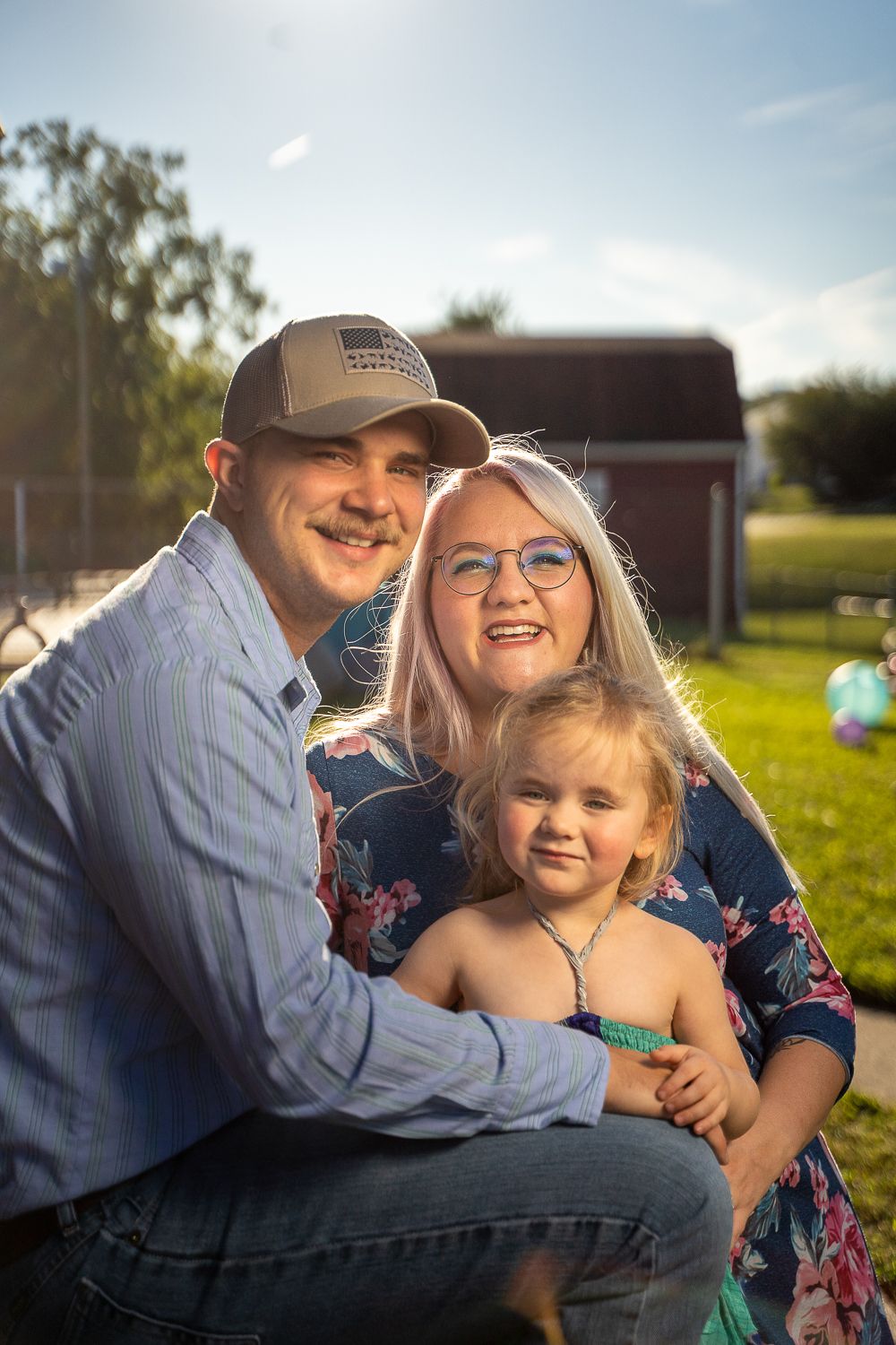 Smiling young family with a toddler posing outdoors in warm sunlight.
