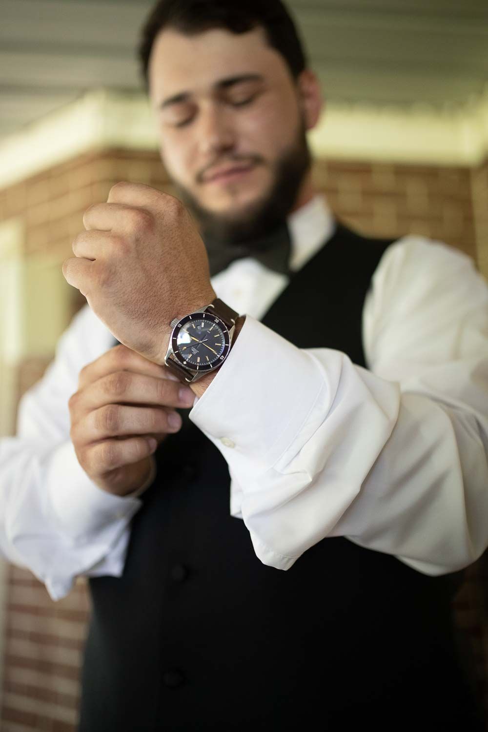 Dapper groom in black vest and bow tie adjusting luxury watch on his wedding day.