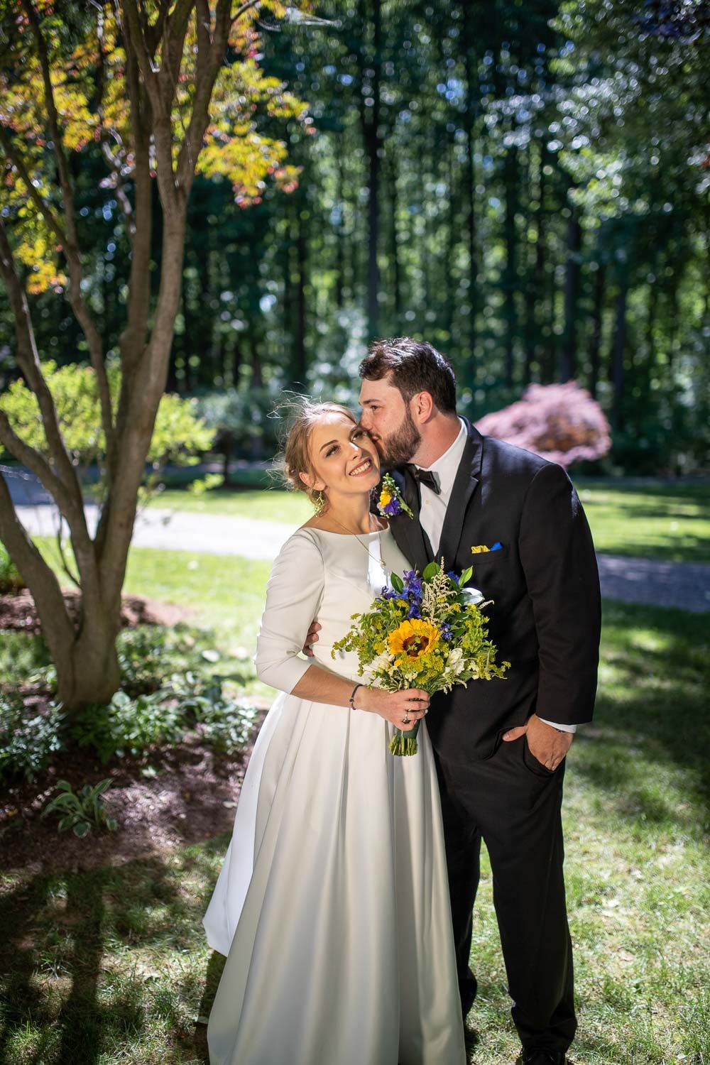 Bride in white satin gown holding sunflower bouquet as groom in black tux kisses her cheek in garden.