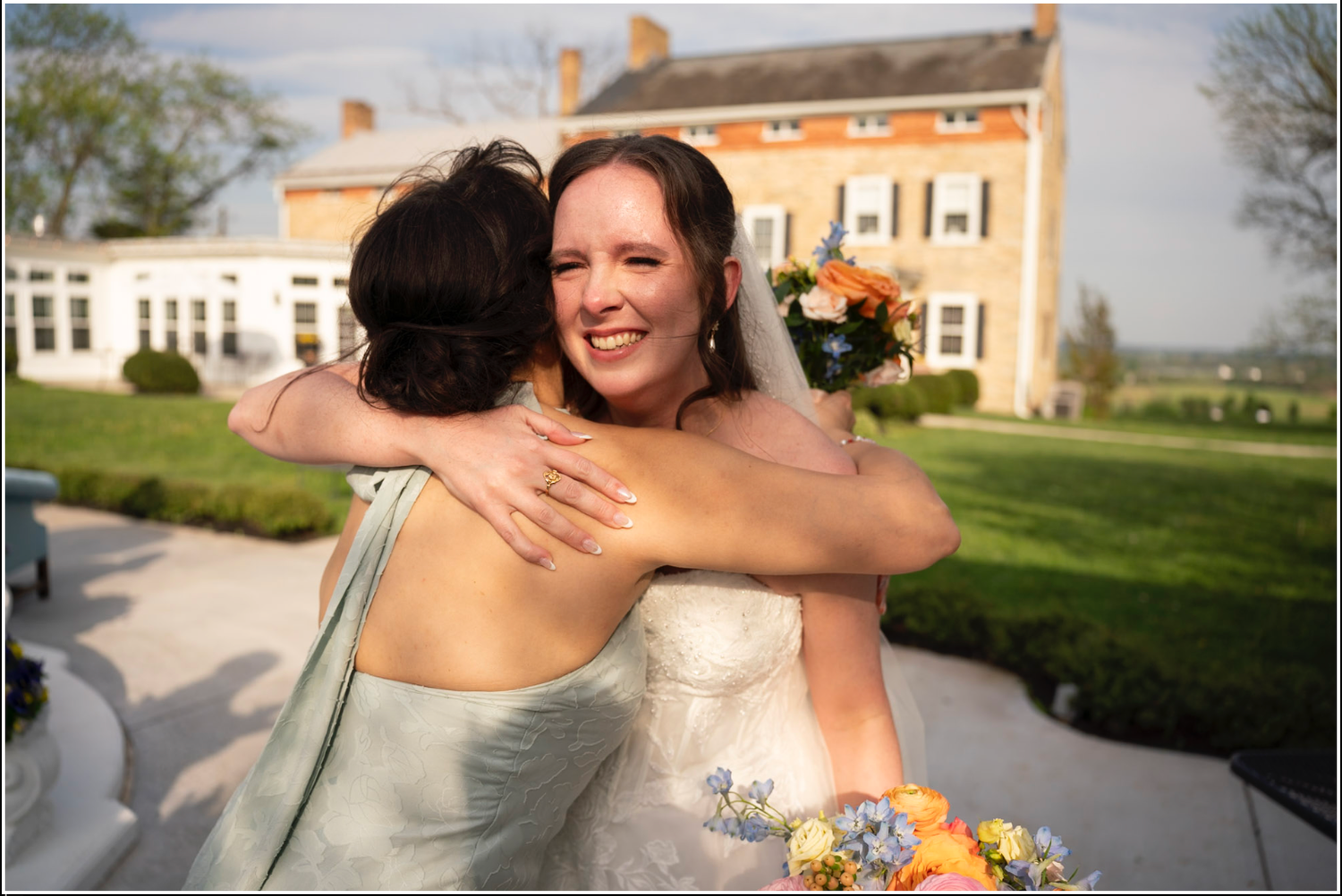 Smiling bride in white gown hugging bridesmaid in blue dress outdoors near elegant brick wedding venue.