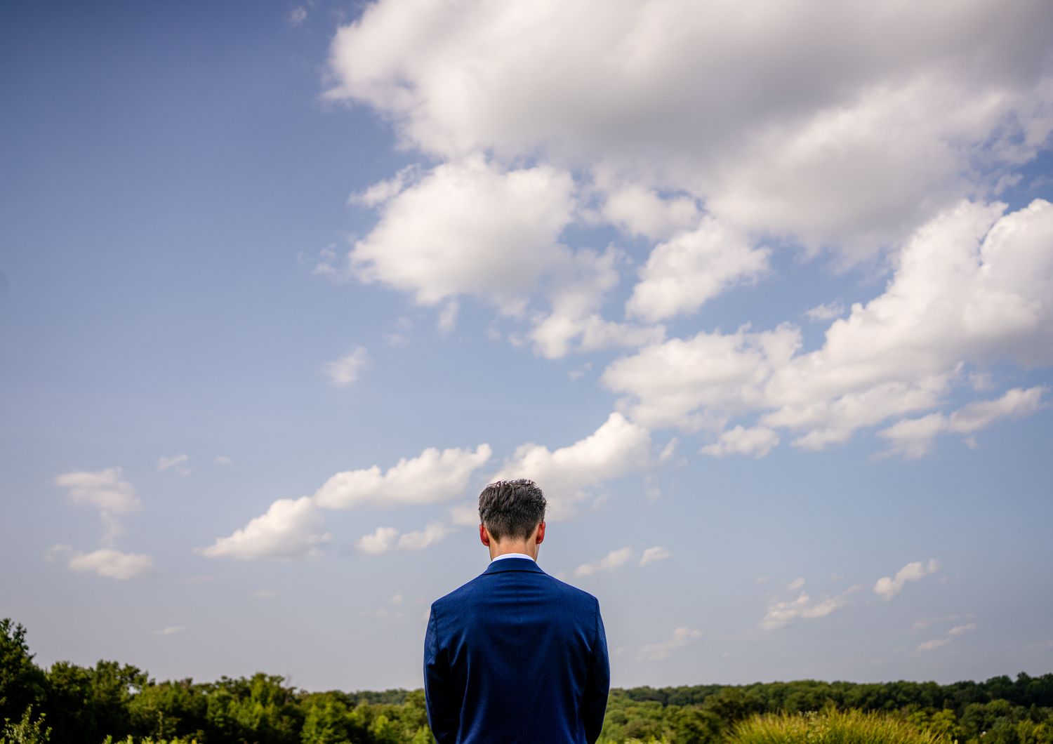 Man in blue suit standing with back to camera gazing at blue sky with white clouds over green trees.
