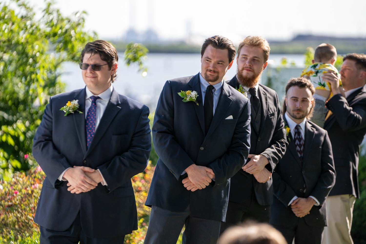 Groomsmen in navy suits with white boutonnieres stand solemnly during outdoor wedding ceremony.