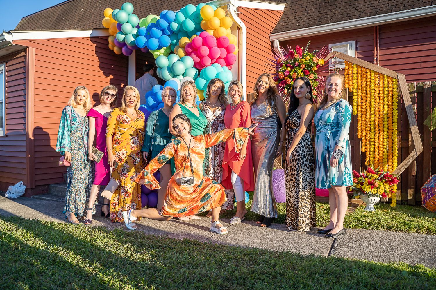 Group of women in vibrant dresses posing together under balloon arch at colorful outdoor party.