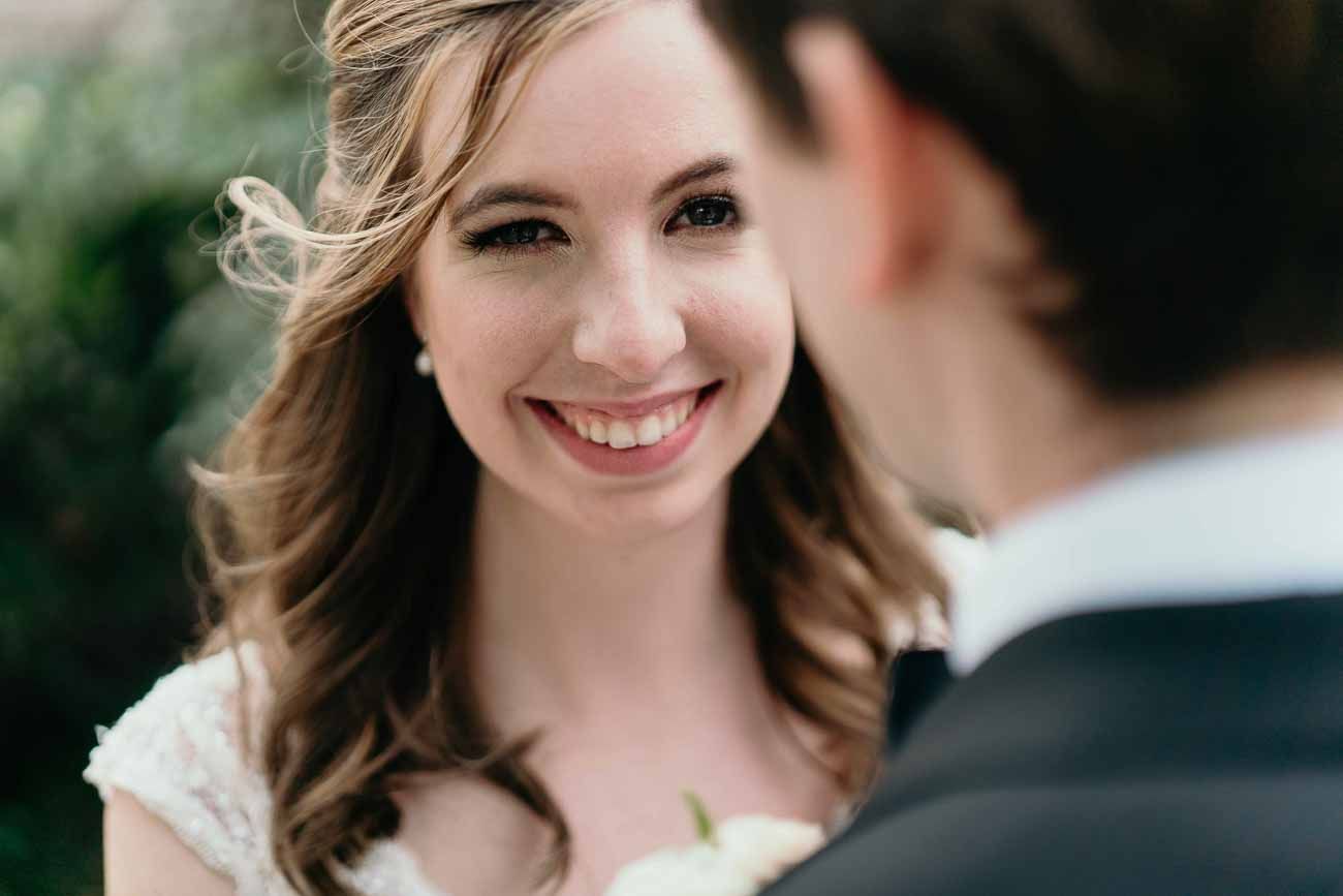 Radiant bride smiles warmly at her groom wearing pearl earrings and soft bridal makeup outdoors.