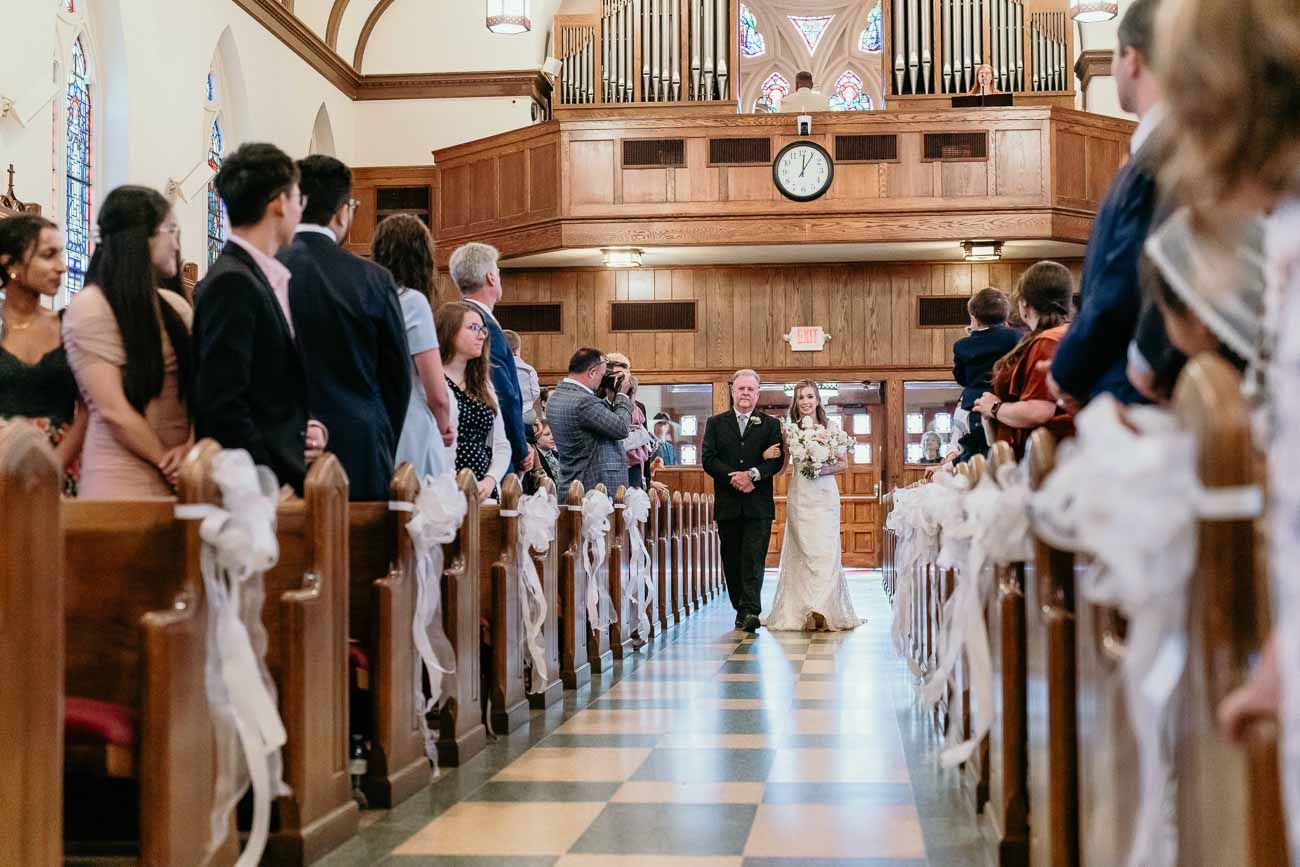 Bride walks down church aisle with escort past guests seated in decorated wooden pews.