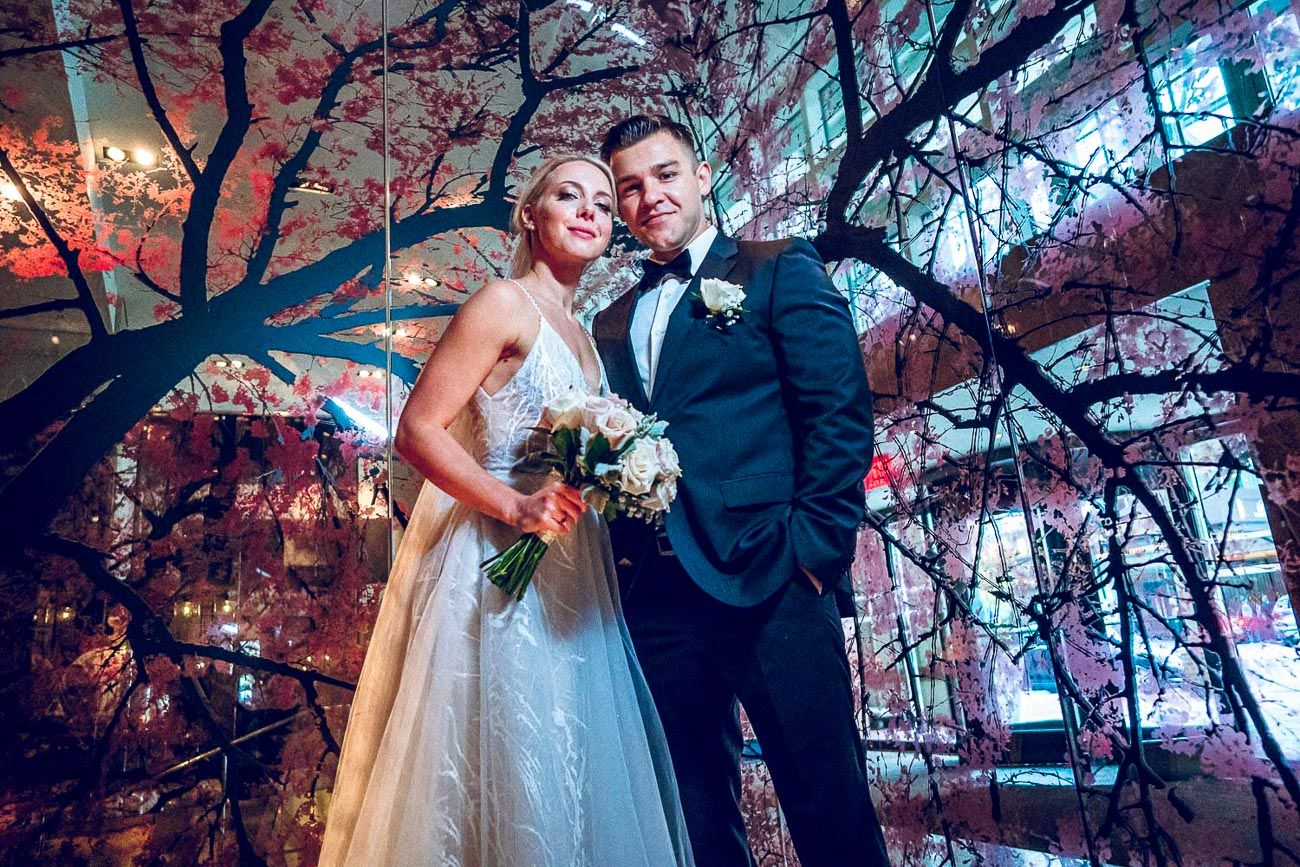 Wedding couple posing elegantly among decorative cherry blossom trees in a vibrant mirrored venue.