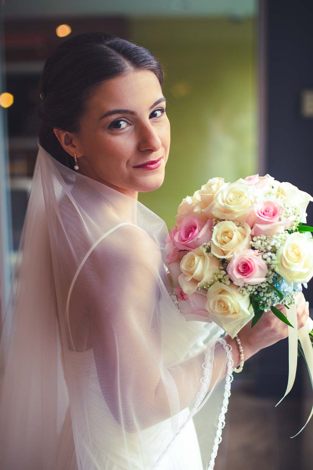 Beautiful bride in white gown and veil holding pink and cream rose bouquet, smiling over her shoulder.