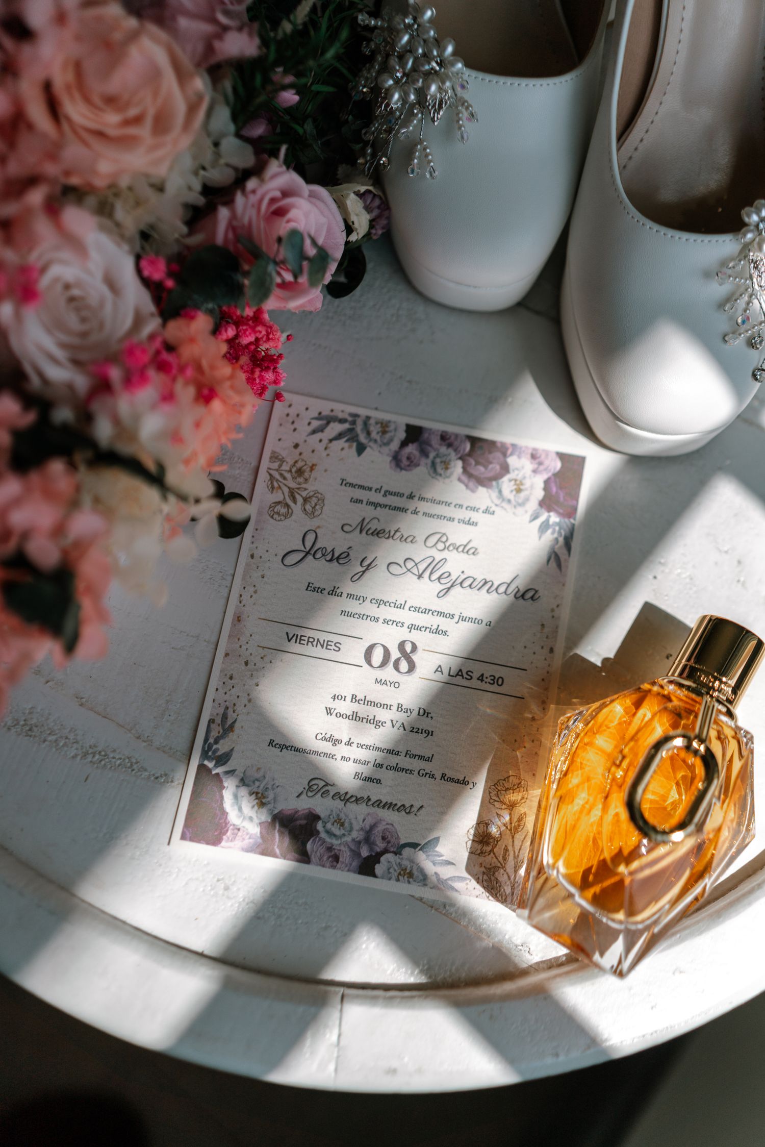 Elegant wedding flat lay with floral invitation for Jose y Alejandra, perfume bottle, white heels, and pink roses on a table.