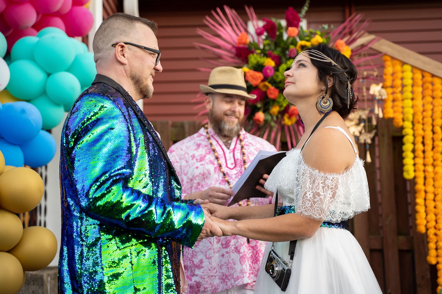 Bride and groom hold hands during colorful outdoor wedding ceremony officiated by man in pink floral shirt.
