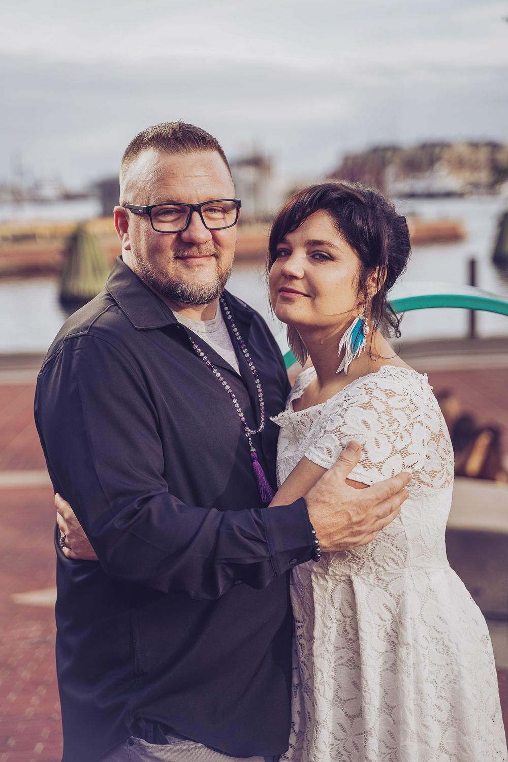 Wedding couple embracing near waterfront harbor, bride in lace dress with turquoise earrings.