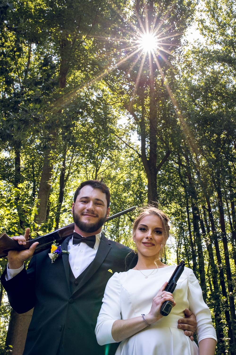 Smiling bride and groom holding shotguns in sunlit forest during outdoor wedding photo shoot.