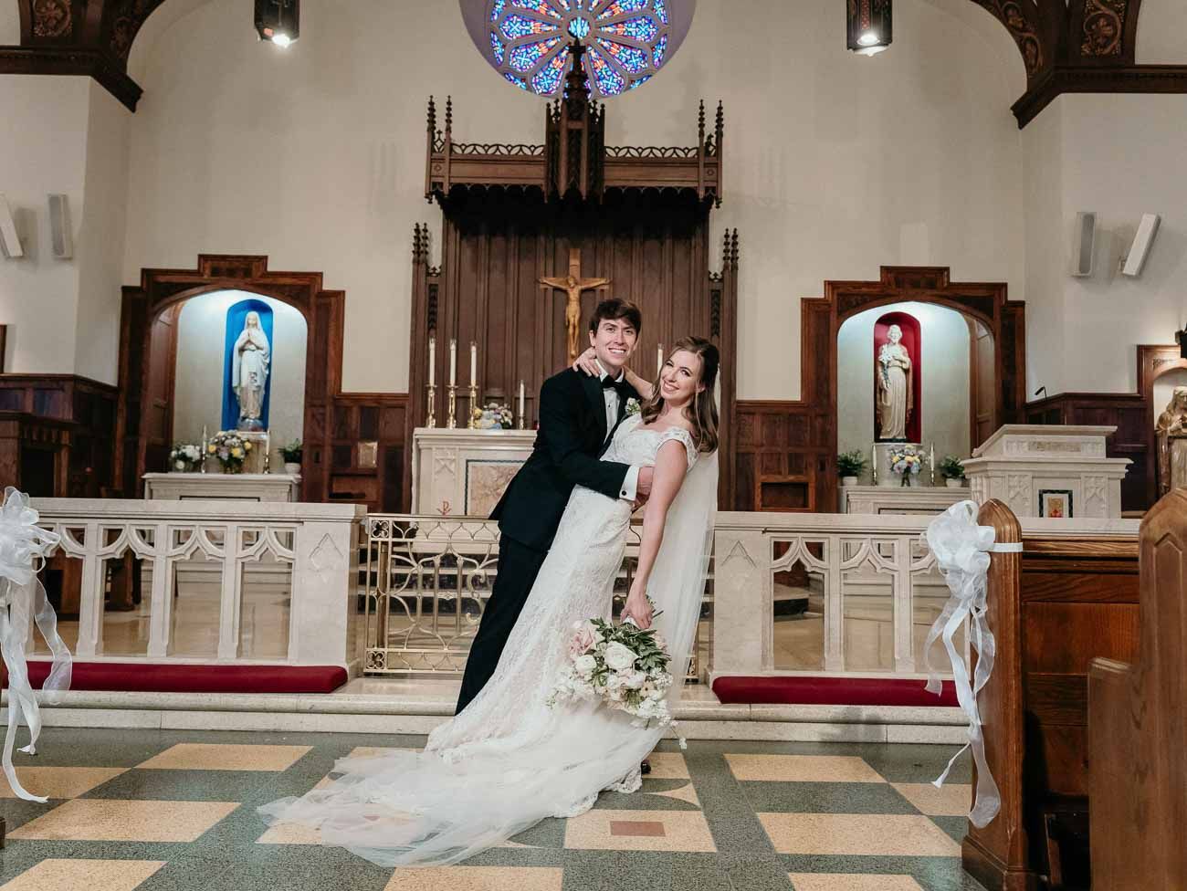 Bride and groom embrace in the aisle of a decorated Catholic church with stained glass windows.