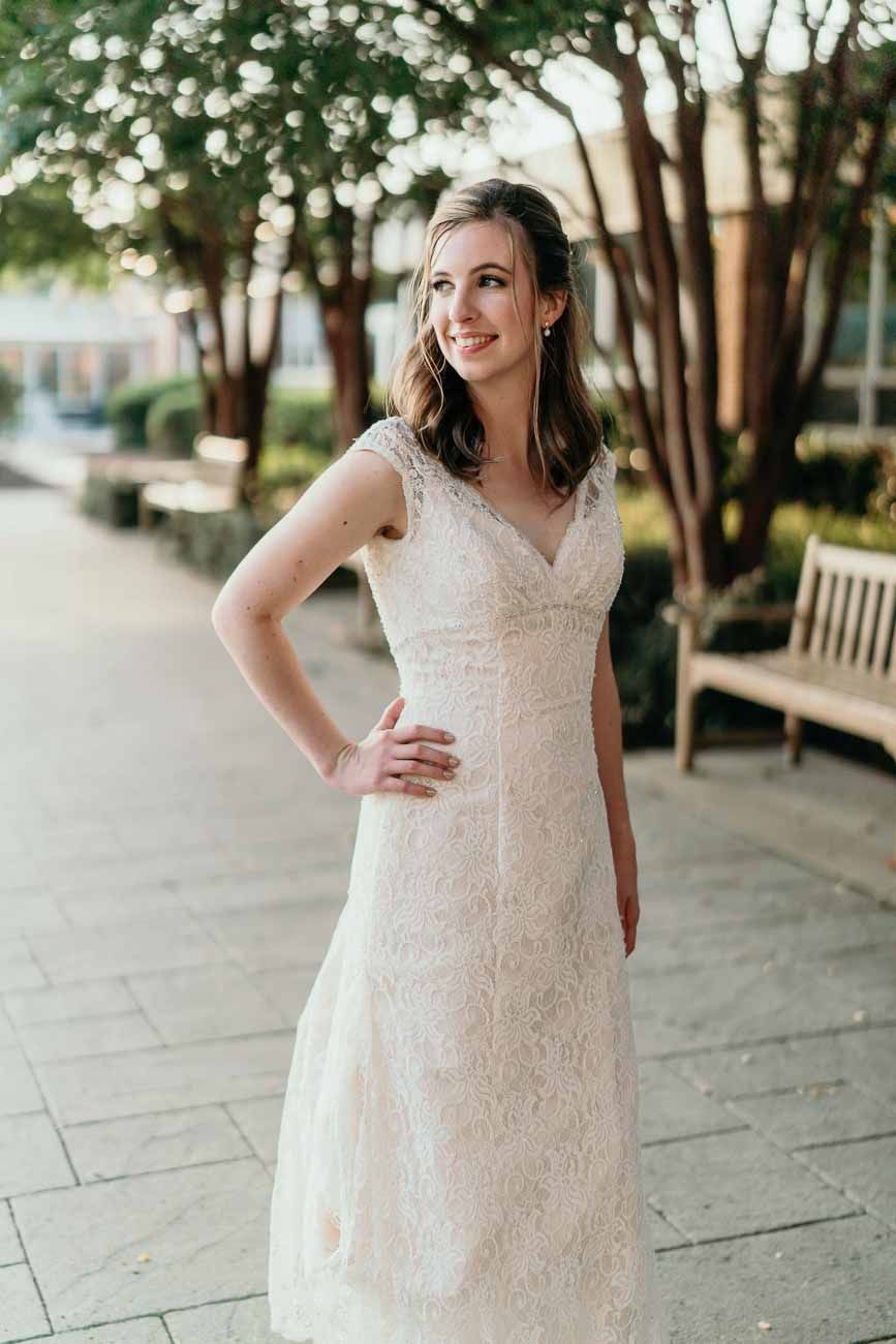 Bride in lace wedding gown poses confidently on outdoor stone pathway surrounded by blooming trees.