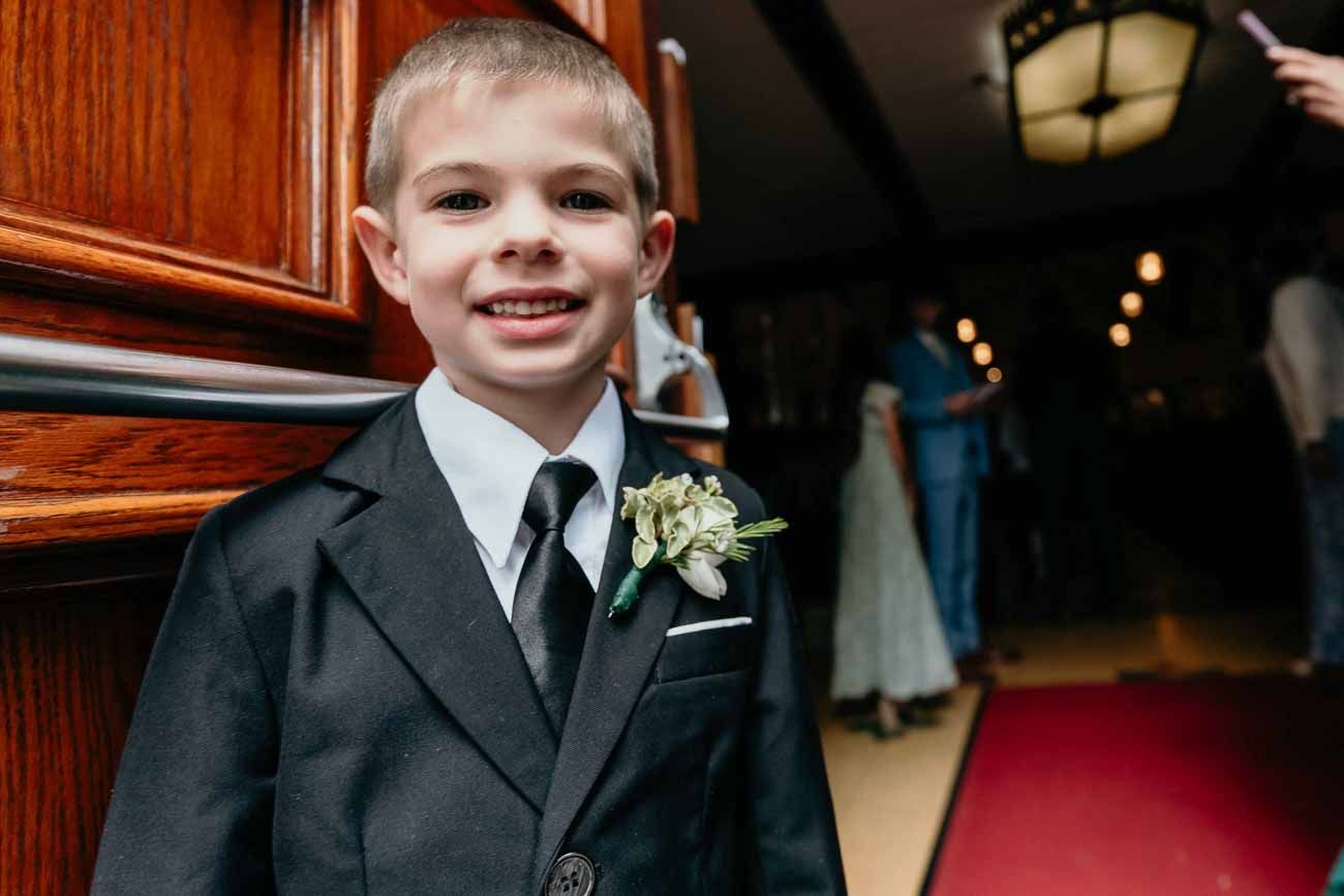 Smiling young ring bearer in black suit with white boutonniere stands by church entrance.