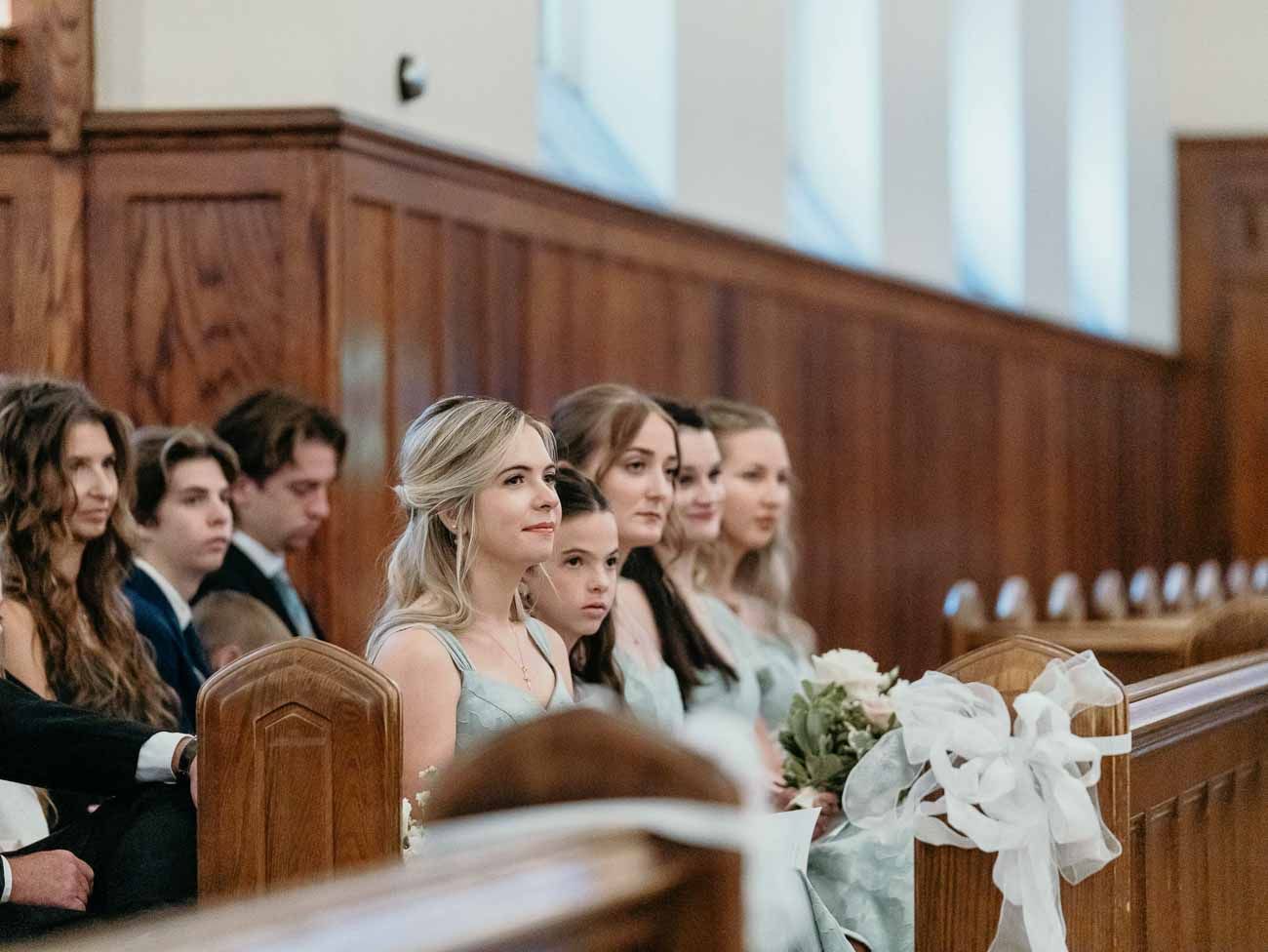 Bridesmaids in sage green dresses seated in wooden church pews during a wedding ceremony.