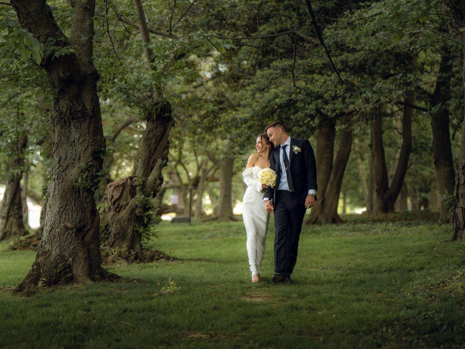 Bride and groom walk hand-in-hand through a lush green wooded area during their wedding day.