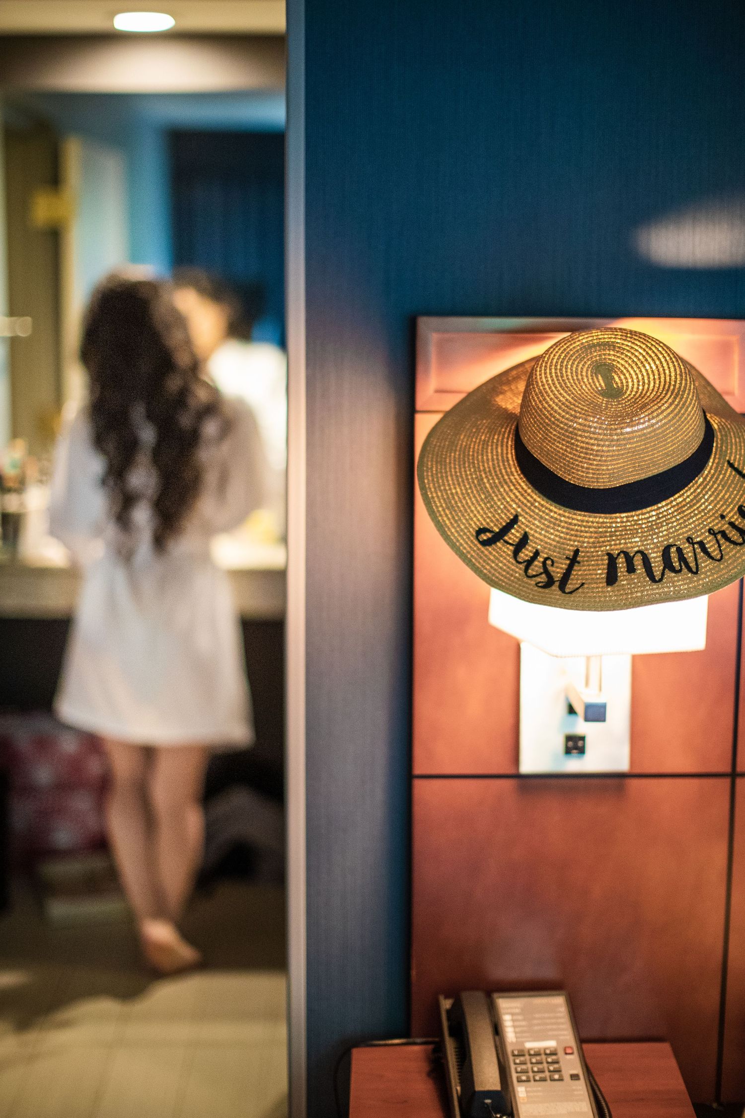 A straw hat reading Just Married hangs on a hotel room wall near a woman in a white robe.