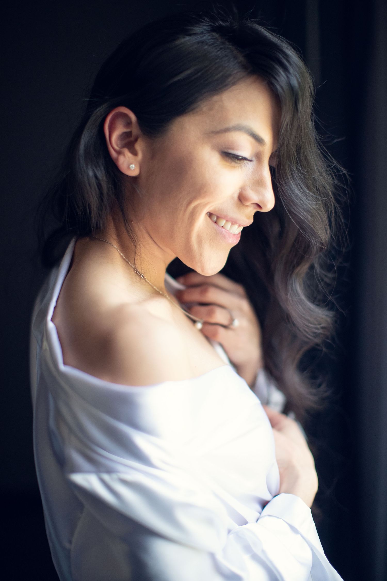Smiling dark-haired bride-to-be in a white off-shoulder robe poses in soft natural light.