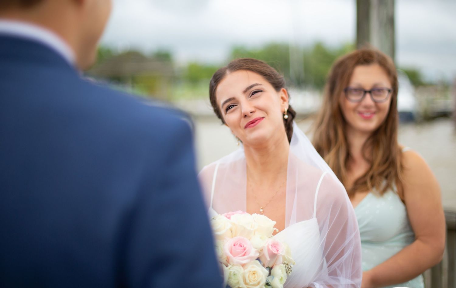 A bride holding a bouquet gazes lovingly at the groom during an outdoor waterfront ceremony.