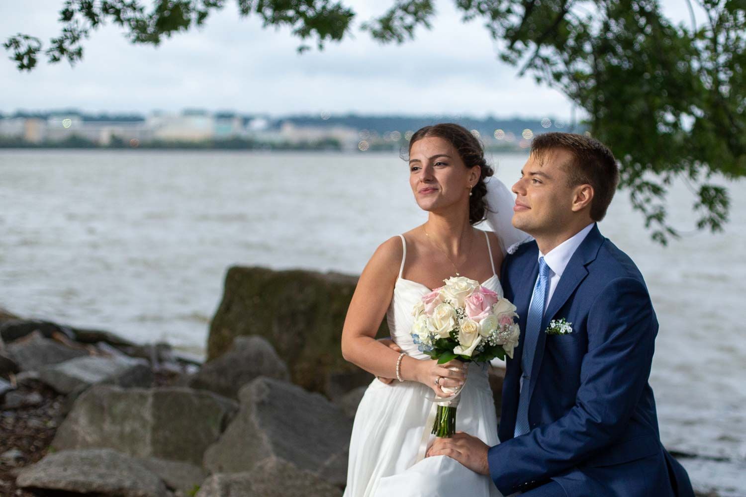 Bride and groom sitting on rocks by the waterfront, bride holding pink and white rose bouquet.