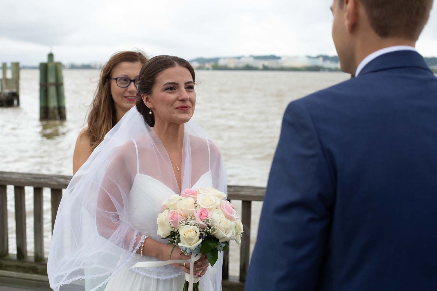 Bride in white gown and veil holding roses gazing at groom during outdoor waterfront ceremony.