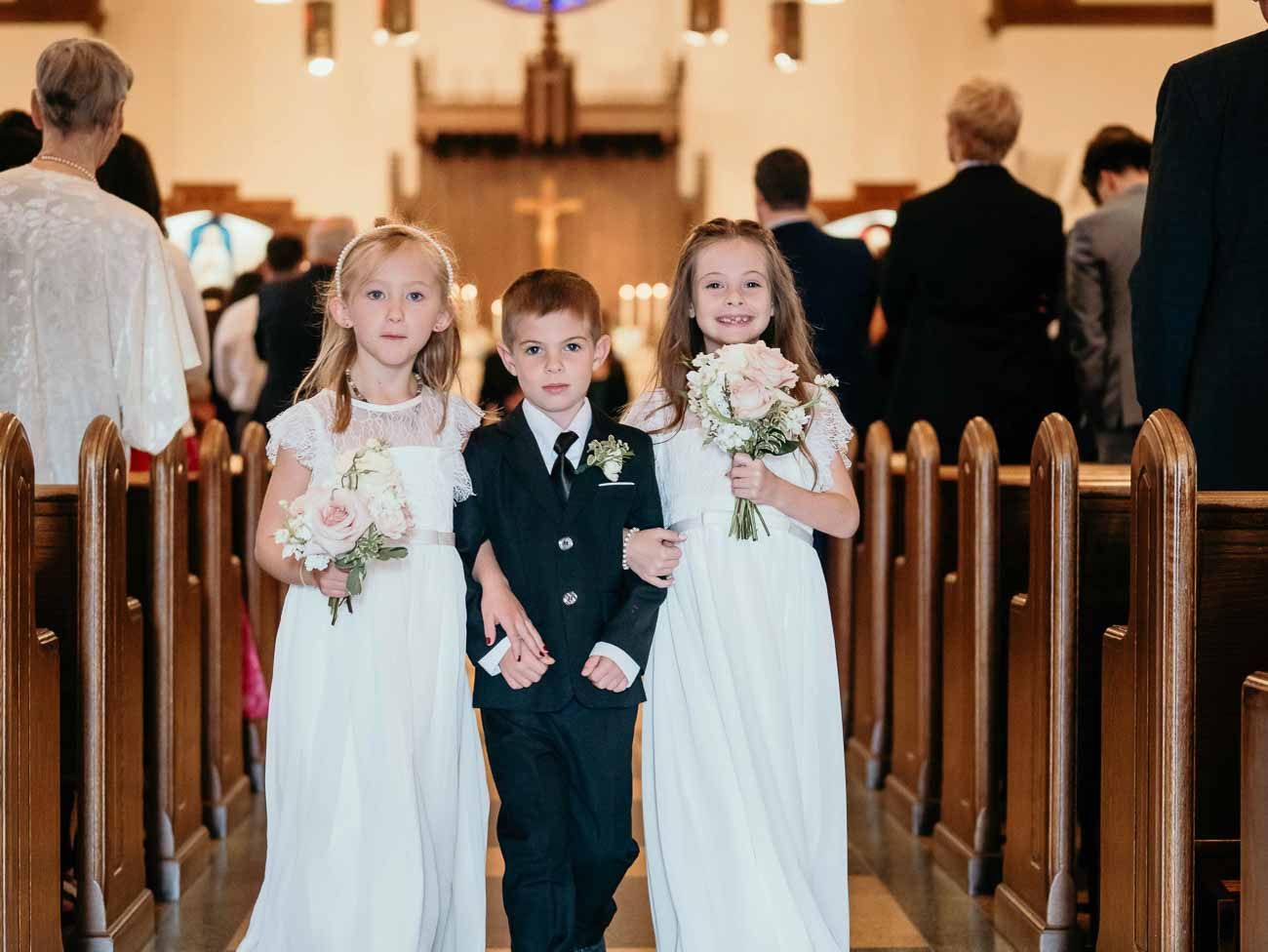Three young children in wedding party pose in church aisle, two flower girls and a ring bearer.