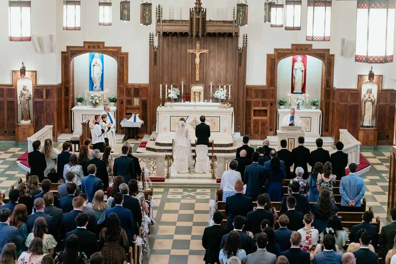 Wide view of Catholic wedding ceremony with bride and groom at the altar surrounded by guests in pews.