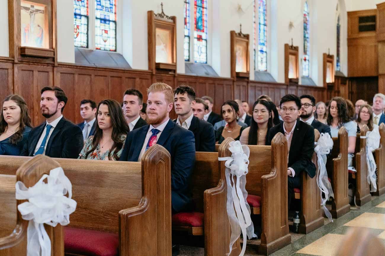 Guests seated in wooden church pews during a wedding ceremony, dressed in formal attire with white ribbon decorations.