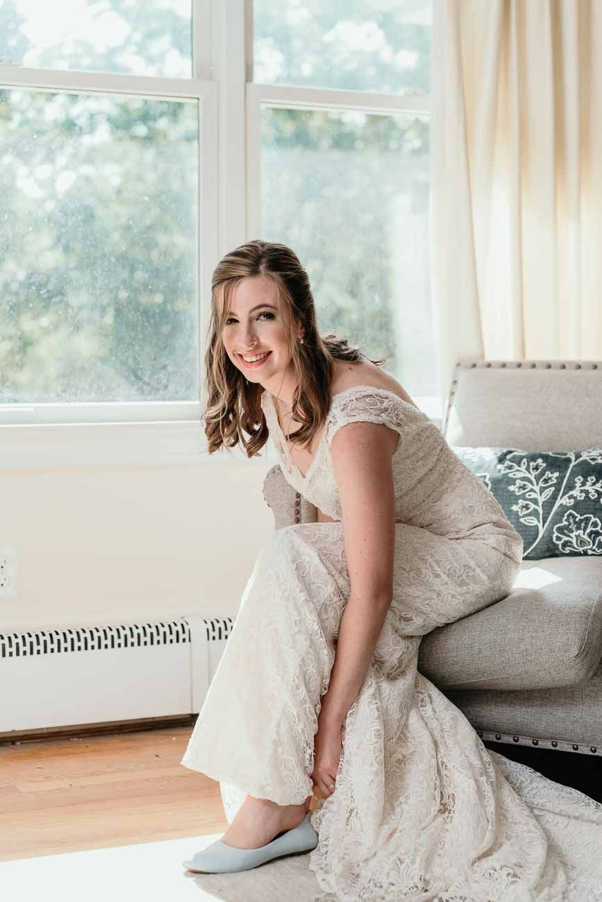 Bride in lace wedding gown smiles while putting on blue flat shoes beside a bright window.
