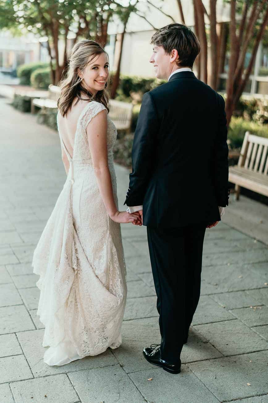 Bride in backless lace gown glances over shoulder while holding groom's hand on a tree-lined walkway.