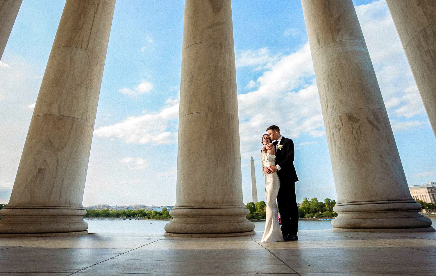 Newlyweds share a tender kiss between towering marble columns at the Jefferson Memorial with Washington Monument view.