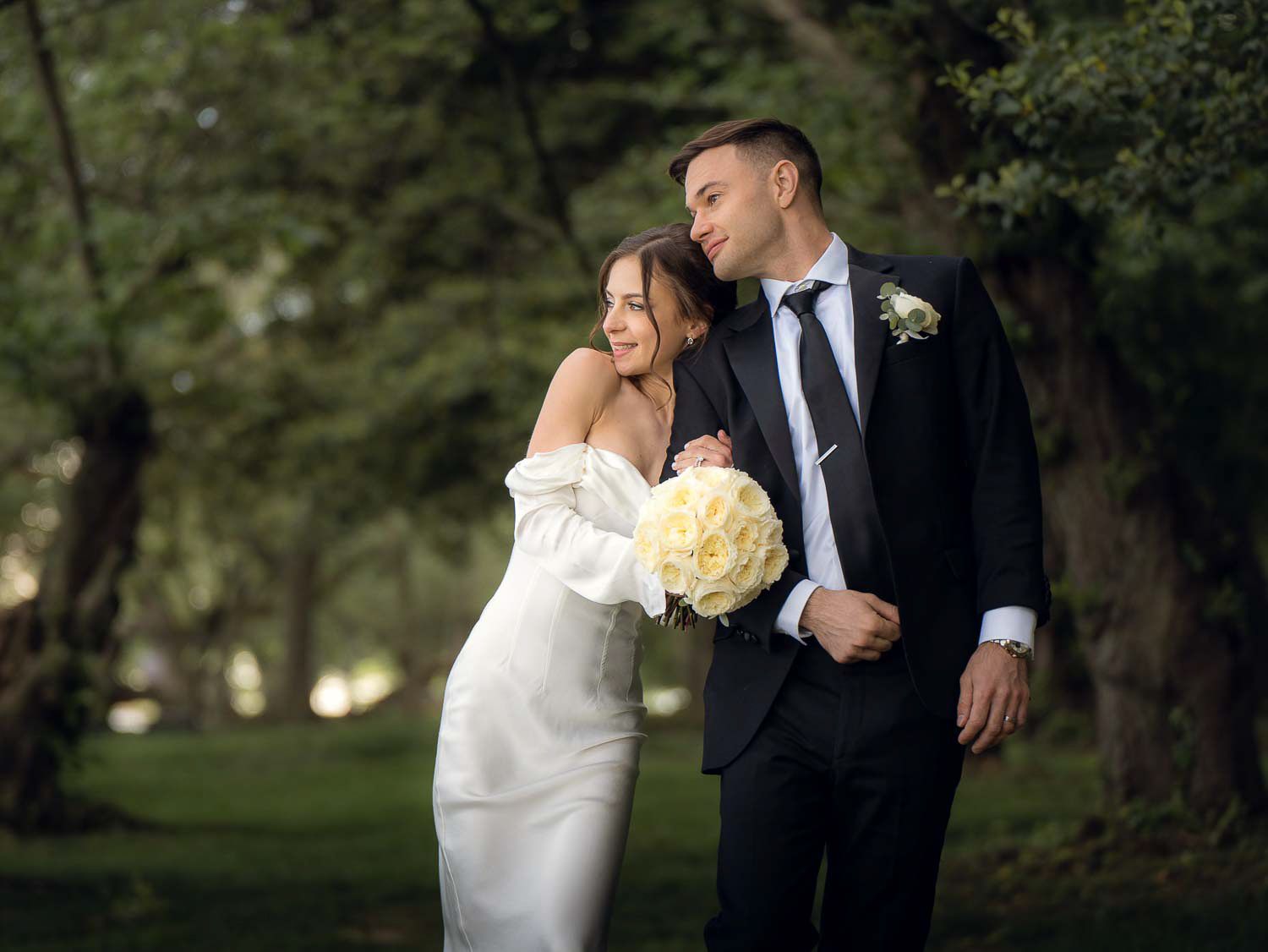 Bride in white off-shoulder gown leans on groom in black suit holding white bouquet in lush green park.