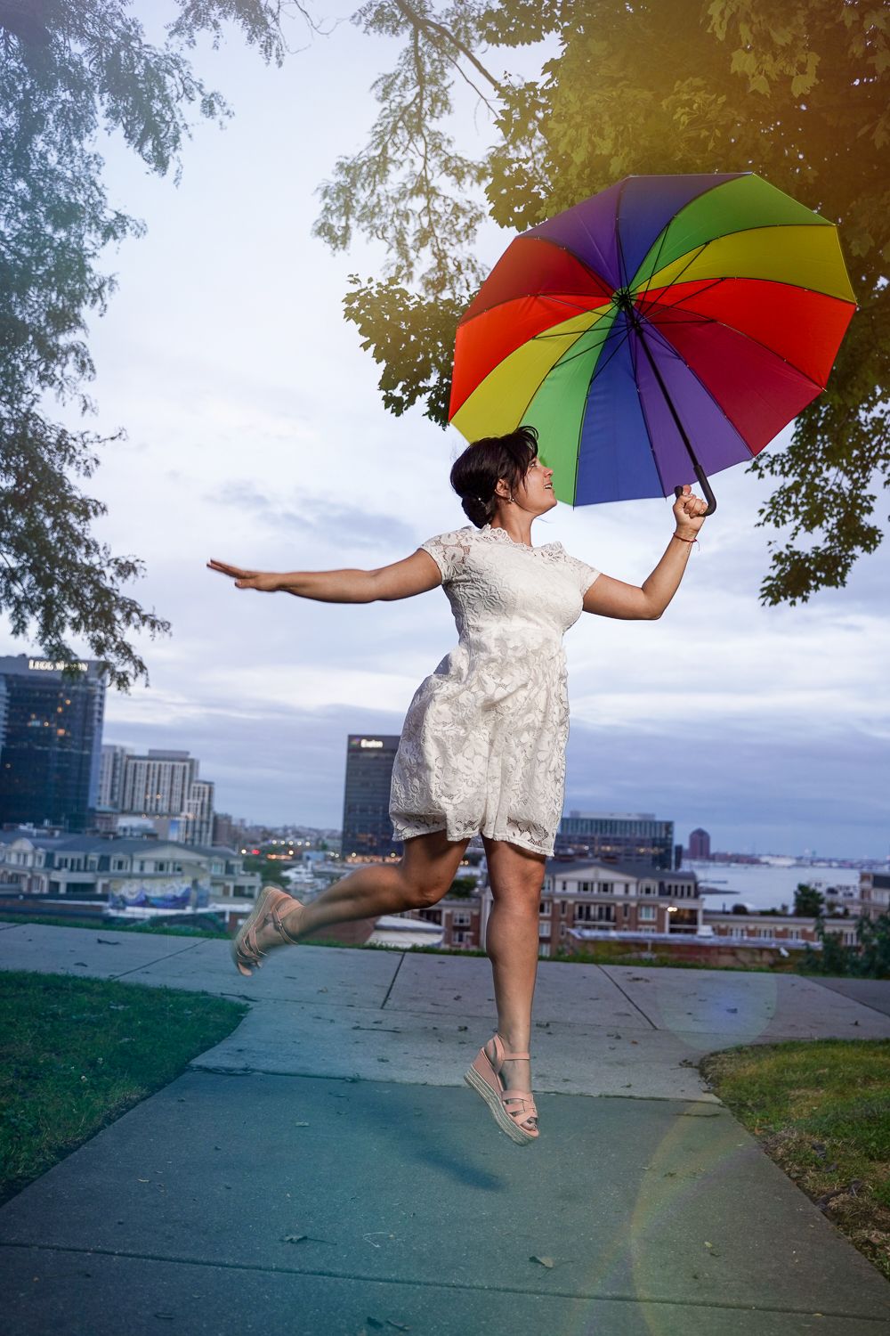 Joyful woman in white dress jumping while holding a vibrant rainbow umbrella with city skyline behind.