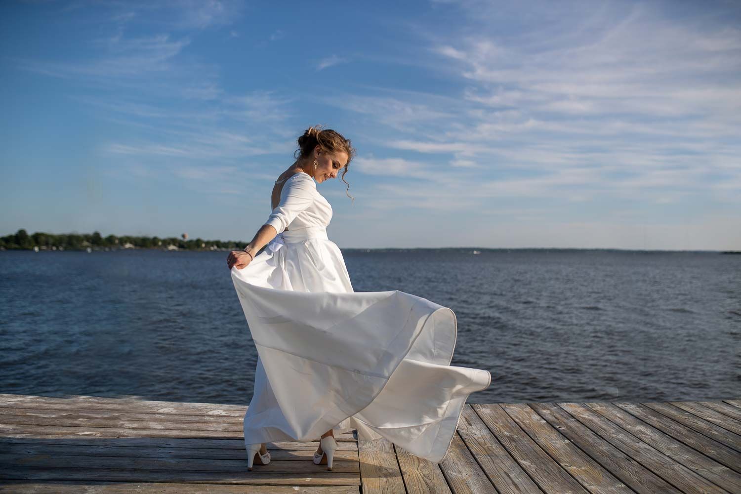 Bride in flowing white gown twirling on wooden dock by the water under blue sky.
