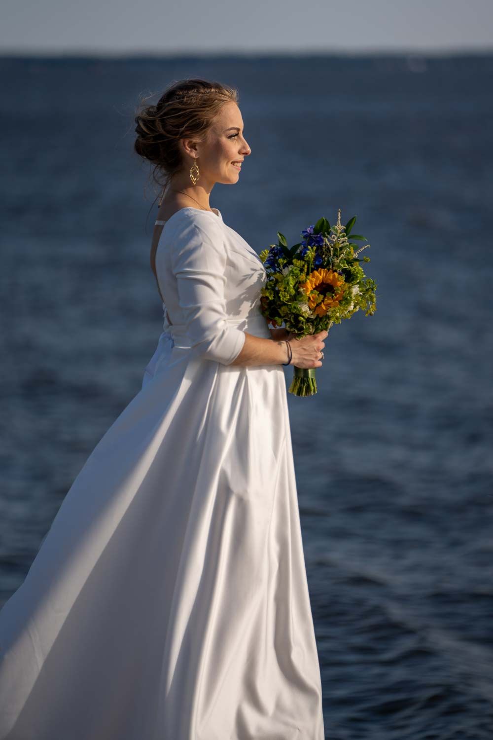 Elegant bride in a white long-sleeve gown holding a sunflower bouquet by the ocean.