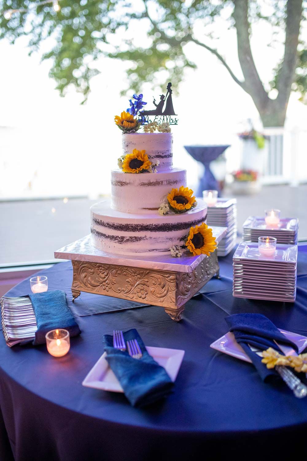 Three-tier white wedding cake decorated with sunflowers on gold stand, topped with silhouette cake topper.