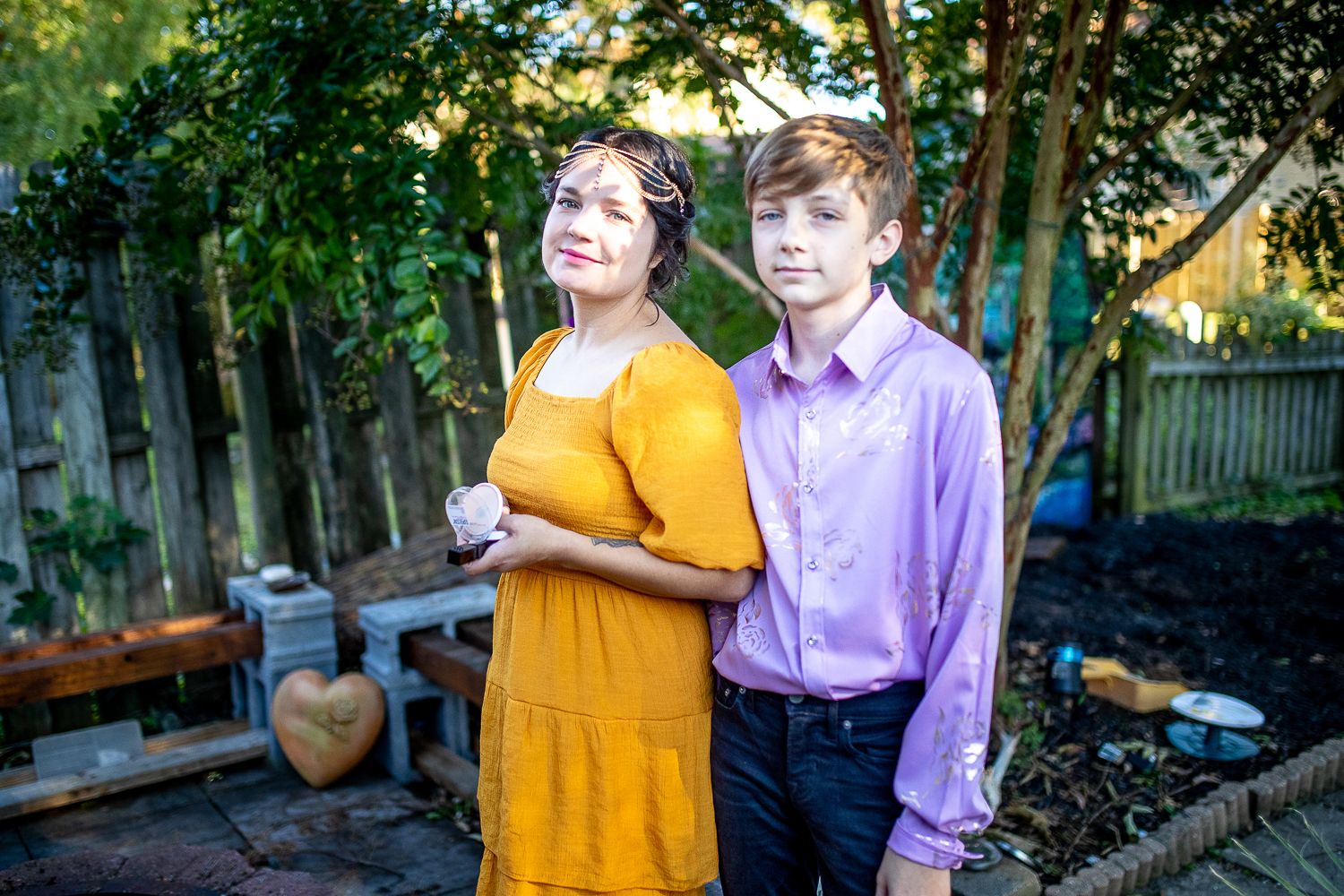 Woman in yellow dress and teen boy in purple shirt posing together in backyard.
