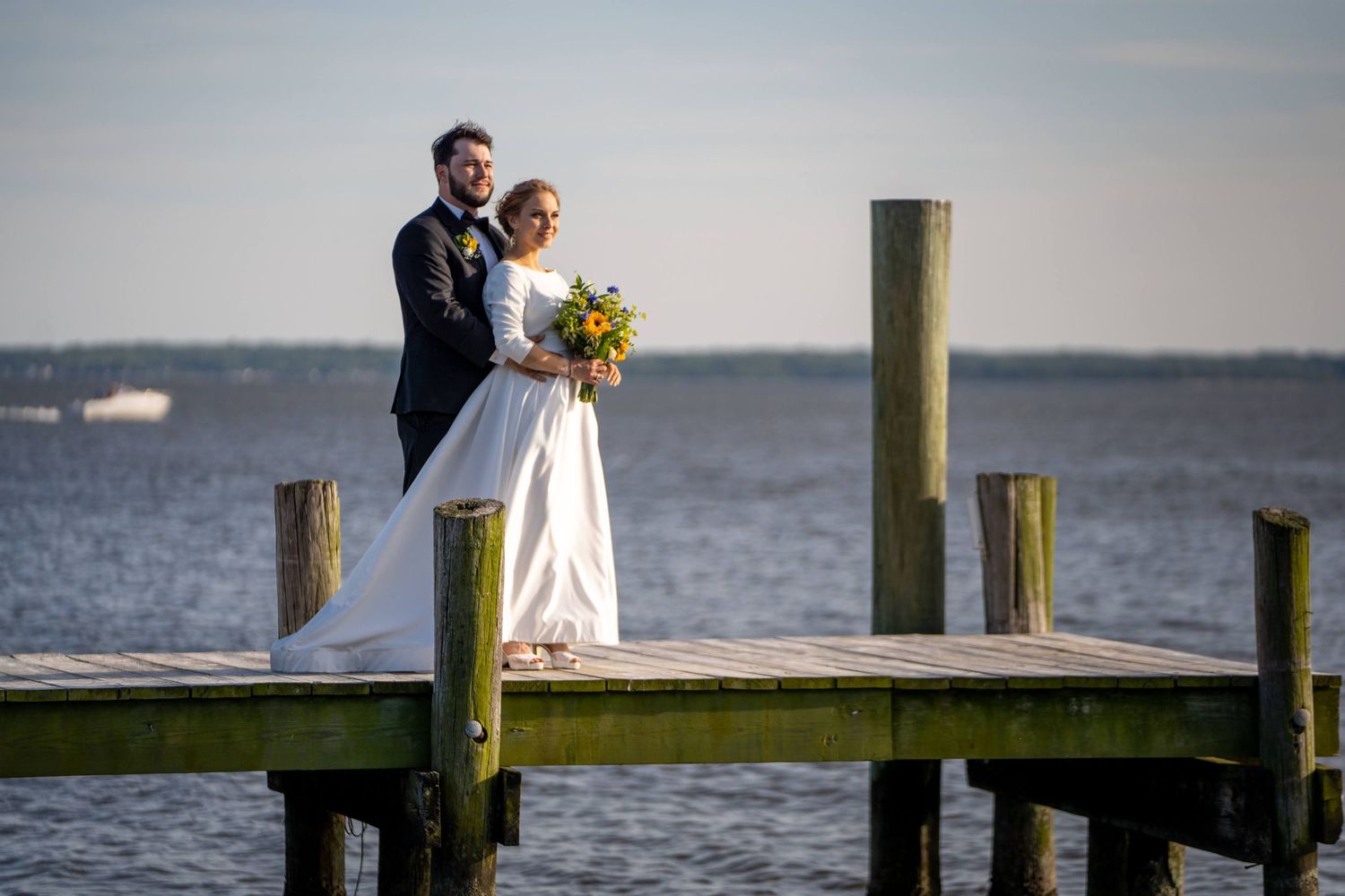 Bride and groom pose romantically on wooden dock overlooking calm water at golden hour.
