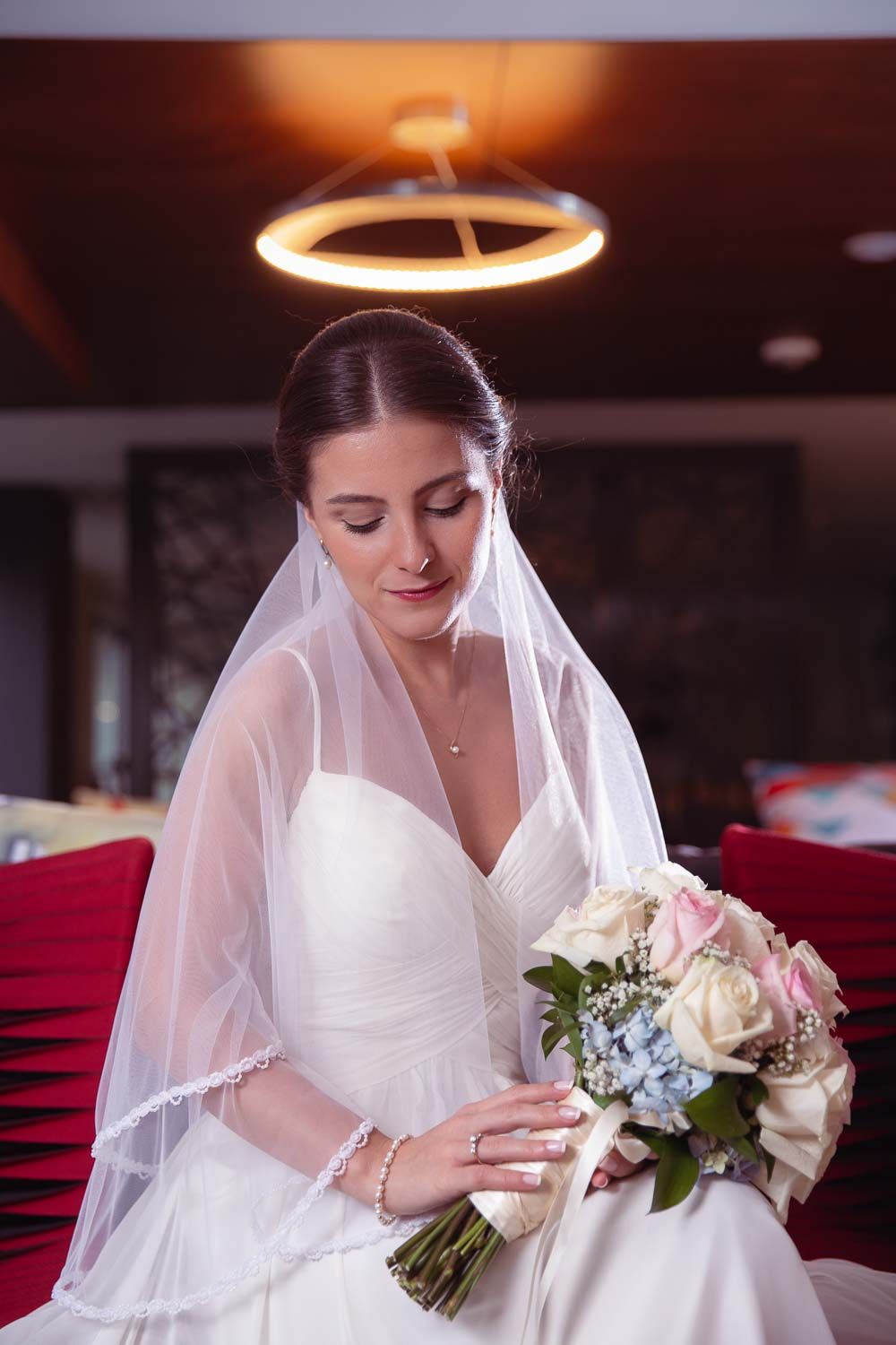 Serene bride in white wedding dress and veil seated holding a pastel rose bouquet under warm lighting.