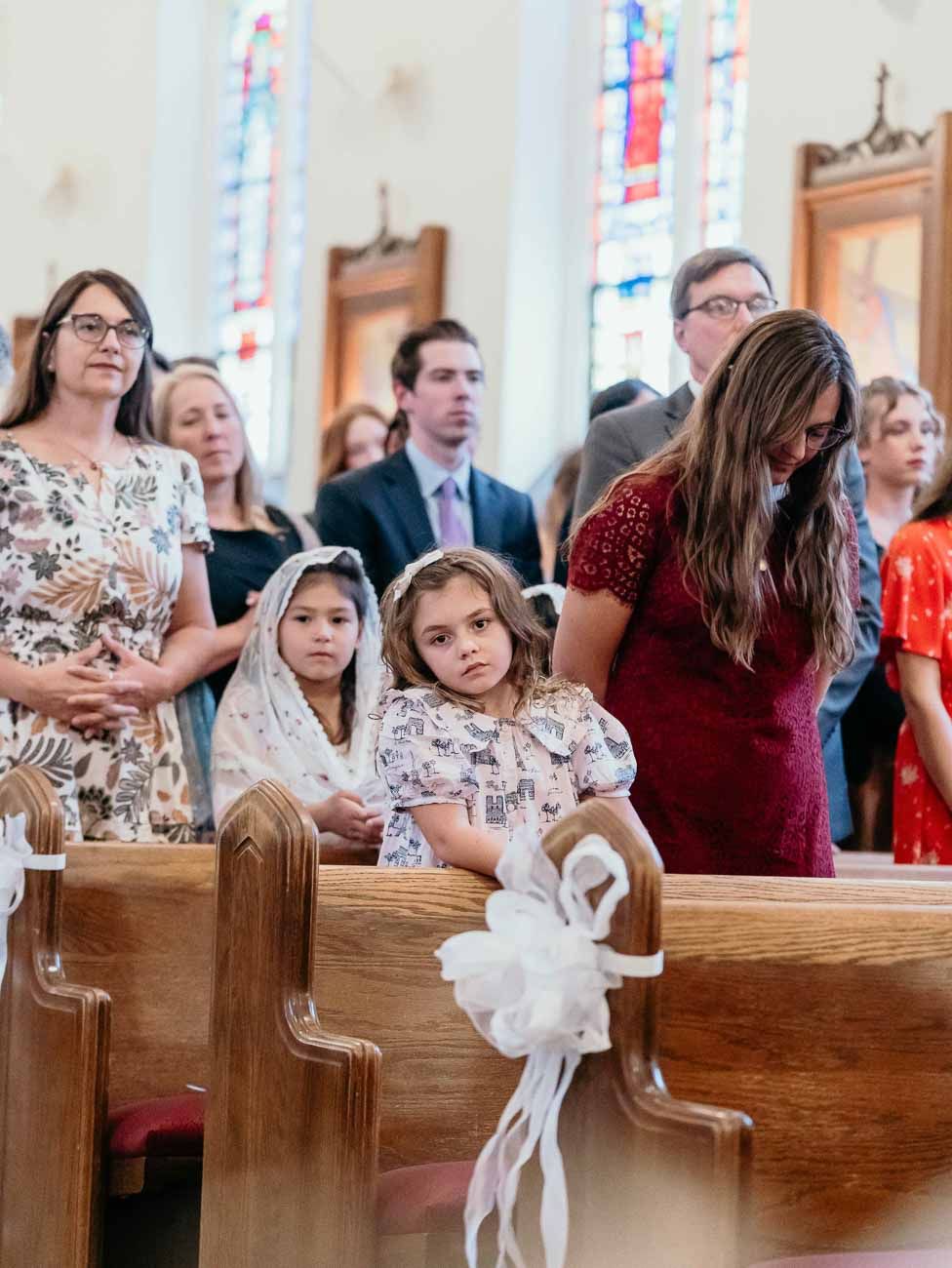 Wedding guests including two flower girls in white veils stand in decorated church pews.