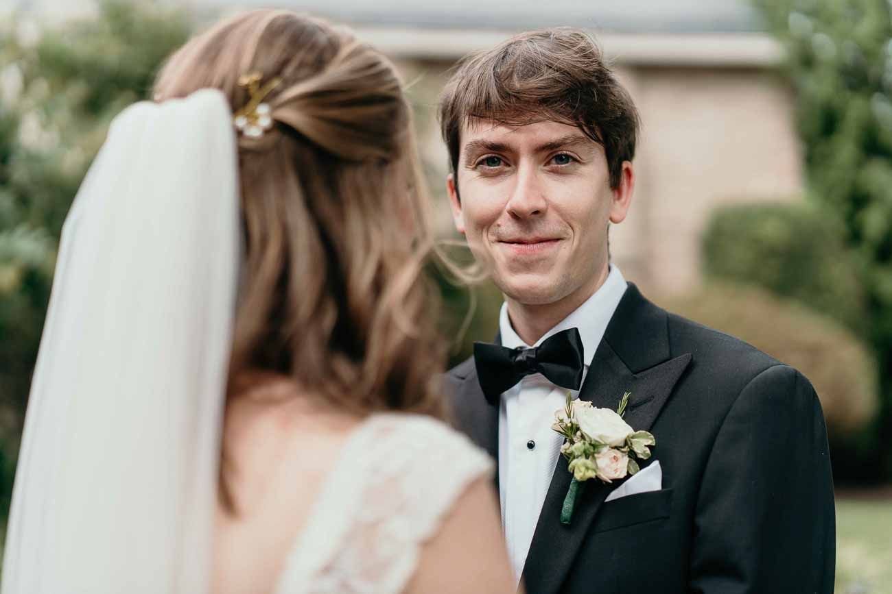 Groom in black tuxedo smiles tenderly at his bride during an outdoor wedding portrait near a church.