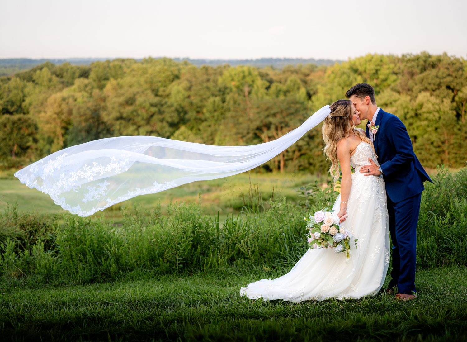 Bride and groom kissing outdoors with flowing lace veil billowing in wind, lush green landscape backdrop.