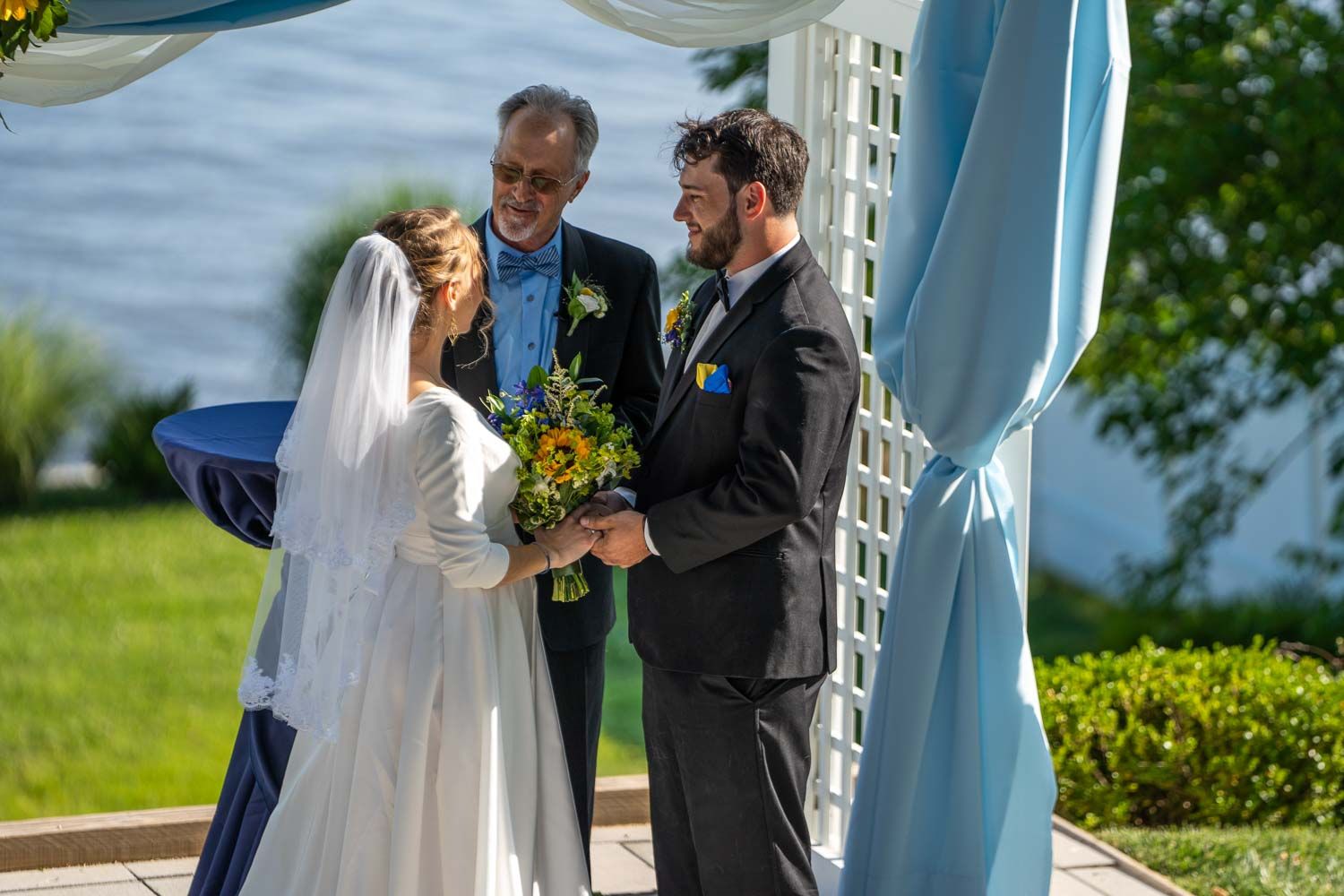 Bride and groom exchange vows with officiant at outdoor waterfront wedding under blue draped arch.