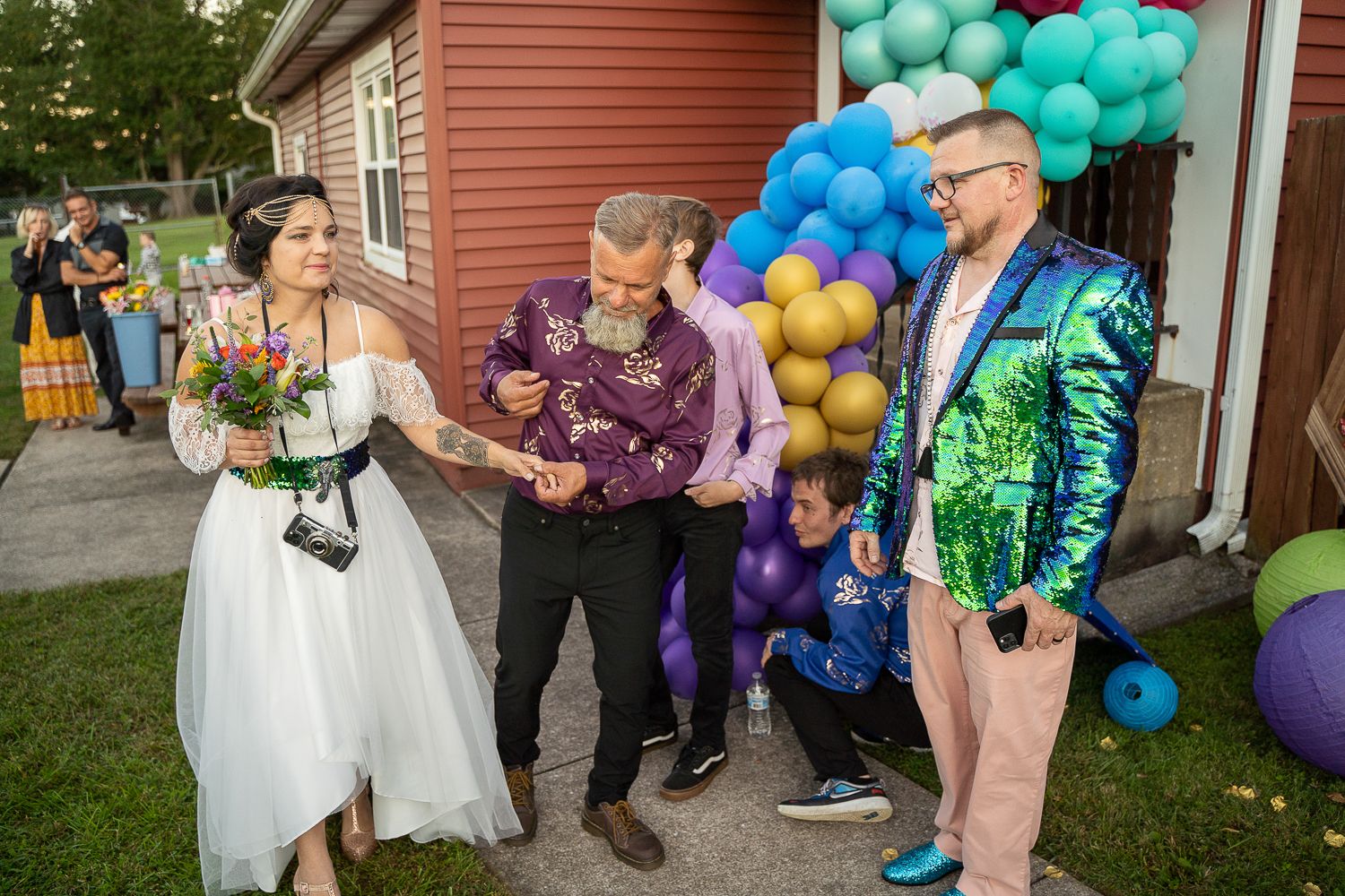 Wedding party gathers outdoors with colorful balloon arch, bride in white dress holds bouquet, guests in vibrant attire.