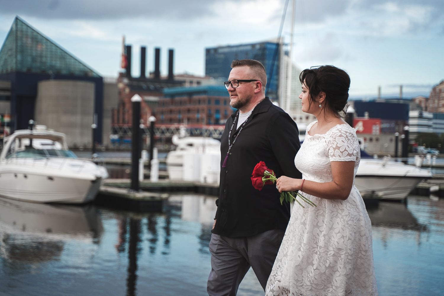 Wedding couple walking along the harbor marina, bride in white lace dress holding red roses, groom in black shirt.