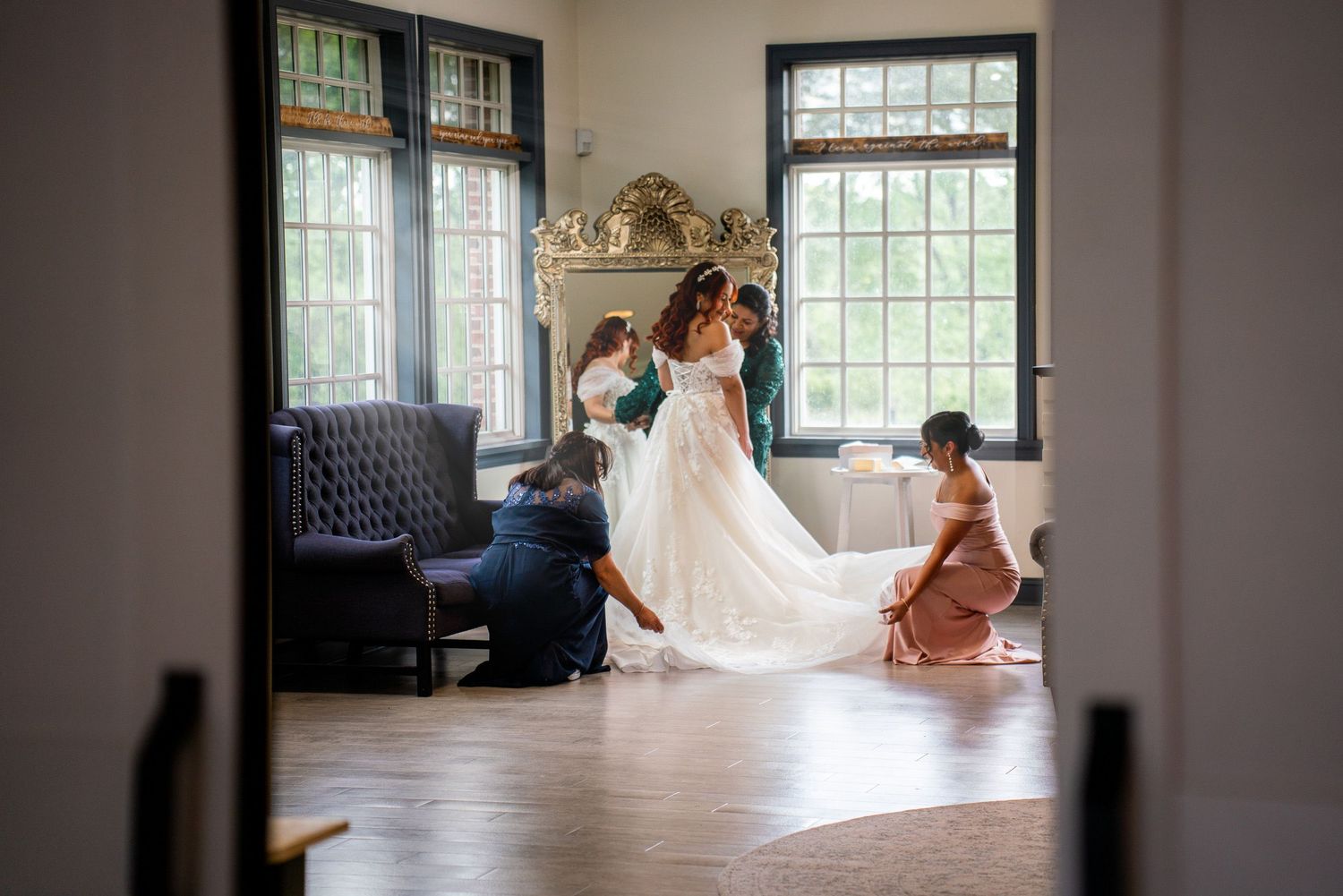 Bride being helped into her wedding gown by bridesmaids in an elegant room with large windows and ornate decor.