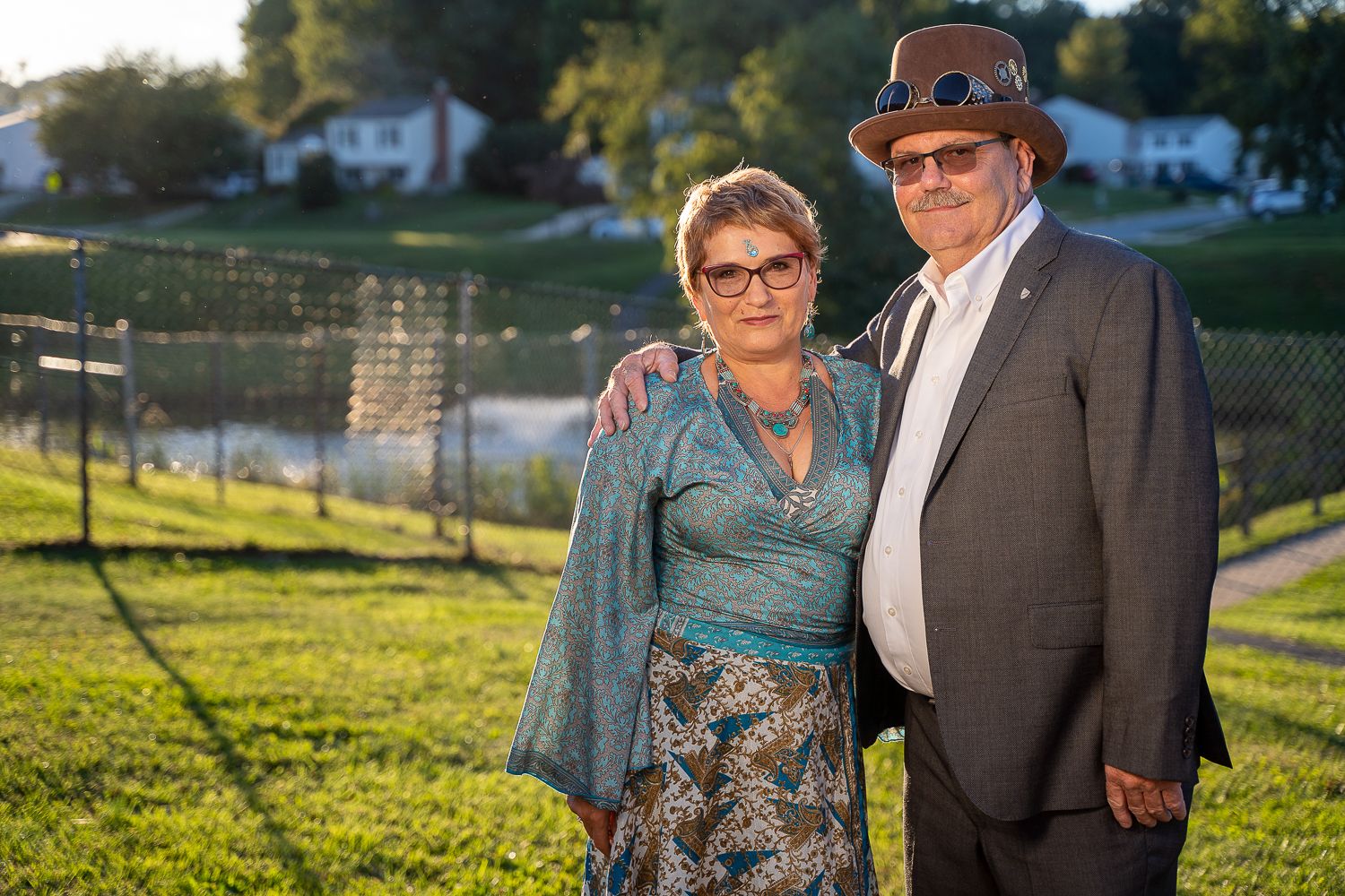 Couple posing together outdoors at golden hour, woman in teal wrap dress and man in hat.